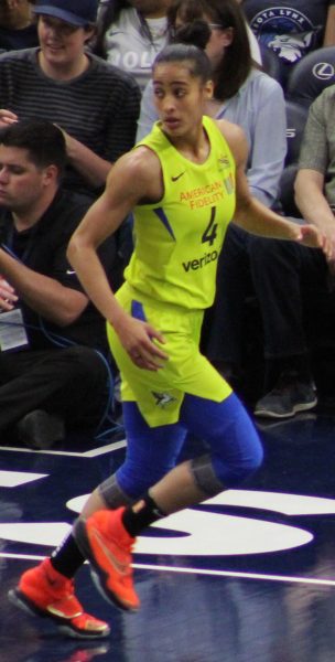 Skylar Diggins running in an electric yellow Dallas Wings uniform with fans seated behind her. 
